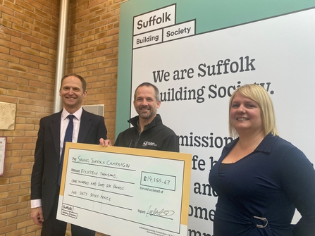 Three people in front of a brick wall with the person in the centre, Michael, holding a giant cheque from the Suffolk Building Society 