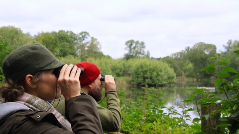 Two people looking through binoculars by a lake